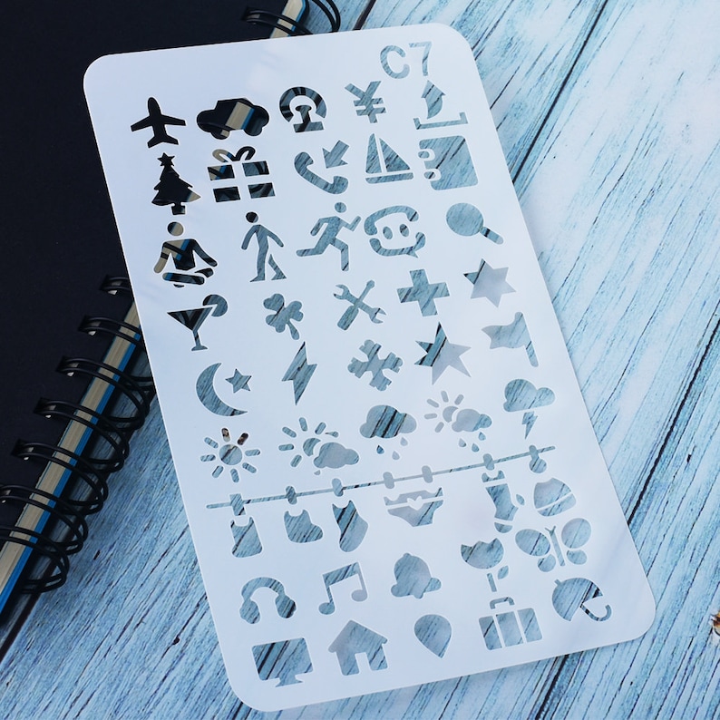 May include: A white plastic stencil featuring cut-out shapes like an aeroplane, a Christmas tree, a gift box, and a musical note. The stencil also has the text "C7". It is placed on a wooden surface. The stencil is for crafts.