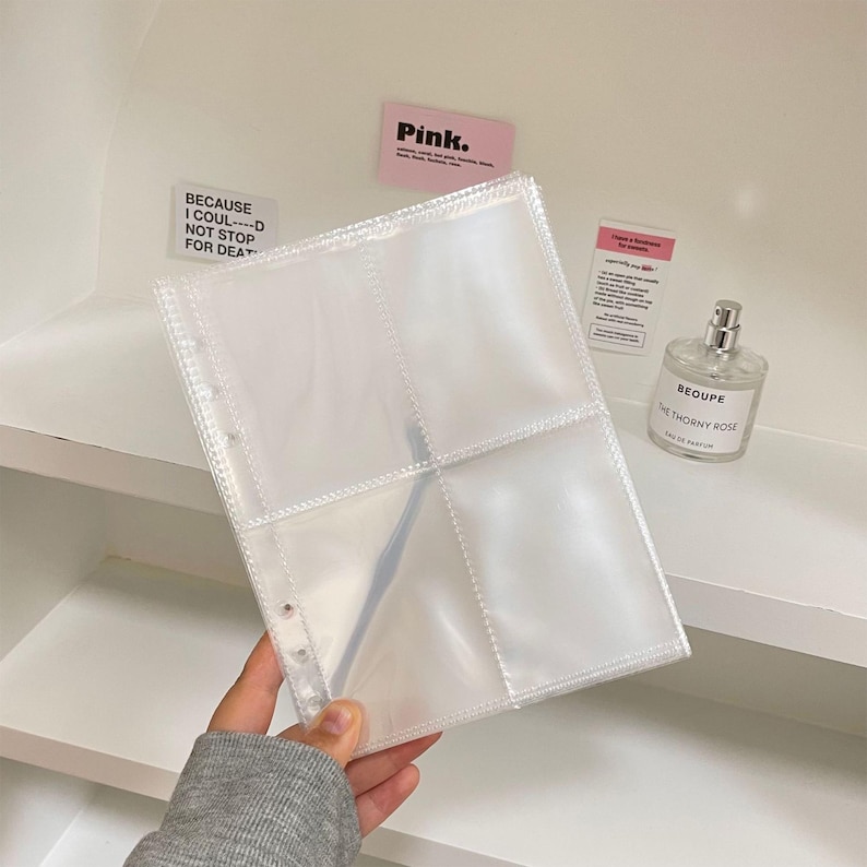 May include: A clear, plastic photo album page with four pockets. The page is held by a person wearing a gray sweatshirt. The background includes a perfume bottle, and two small pink and white signs. One sign reads "Pink."