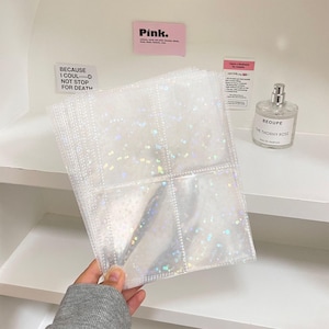 May include: Clear, sparkly photo album pages with multiple pockets. The pages are held in a hand, and the pockets are designed to hold small photos or cards. The background includes a white shelf and a perfume bottle.