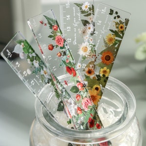 May include: Four clear plastic rulers, each 15 cm long, with floral designs of roses, daisies, and sunflowers. The rulers are in a glass jar. The text "Dream as if you'll live forever" is visible on one ruler.