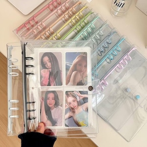 Puede incluir: Un archivador de plástico transparente con anillos de colores arcoíris. El archivador está abierto y tiene insertos para tarjetas de fotos con imágenes de cuatro mujeres. El archivador está sobre una superficie blanca.