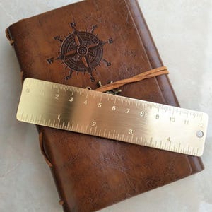 May include: A brass ruler, 12 inches long, rests on a brown leather journal with a compass rose design. The ruler has both inch and centimeter markings. The journal is closed with a leather strap.