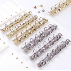 May include: Close-up of several gold and silver binder rings, with a white background. The rings are arranged in rows, with some loose hardware pieces scattered nearby. The rings are designed for use in notebooks or planners.