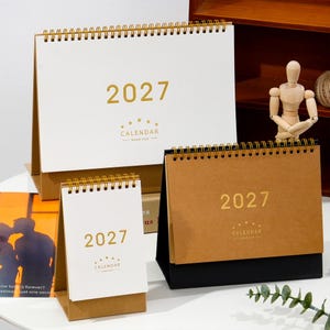 May include: Three desk calendars for the year 2027, featuring gold foil text and spiral binding. One large landscape calendar with a white cover, one small portrait calendar with a white cover, and one medium landscape calendar with a brown cover.