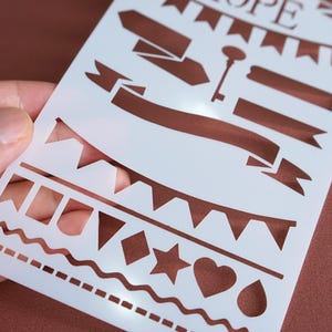 May include: A white stencil featuring various decorative shapes and banners. The stencil includes a key, banners, pennants, a star, a heart, a diamond, and a teardrop. The word "HOPE" is at the top of the stencil.