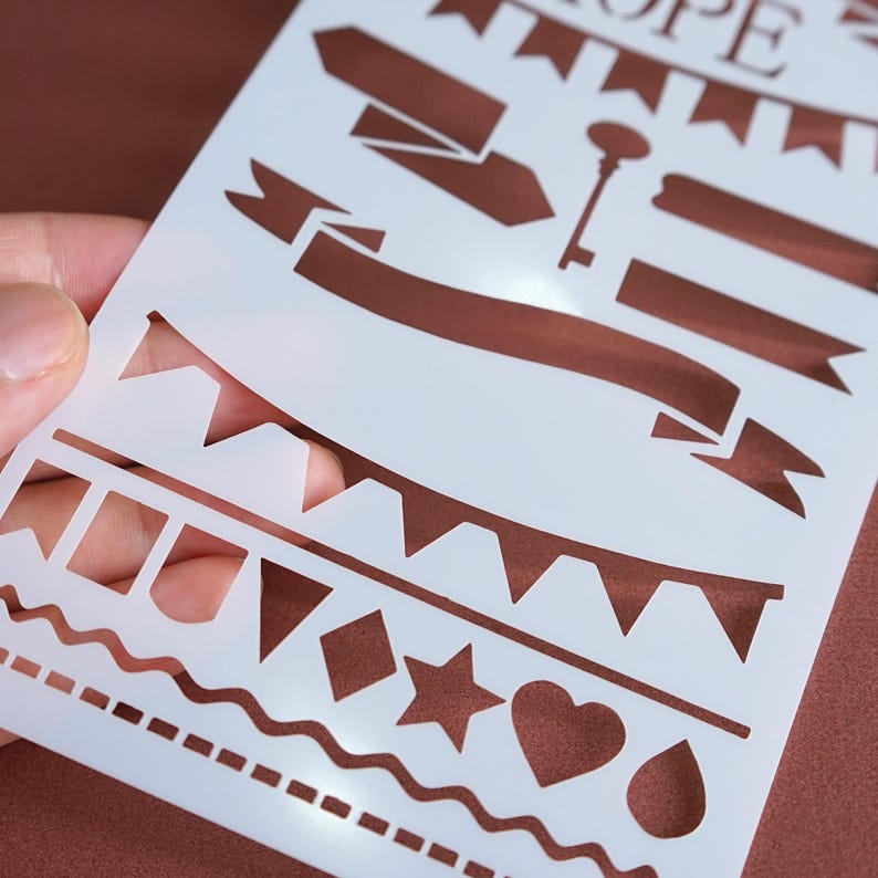 May include: A white stencil featuring various decorative shapes and banners. The stencil includes a key, banners, pennants, a star, a heart, a diamond, and a teardrop. The word "HOPE" is at the top of the stencil.