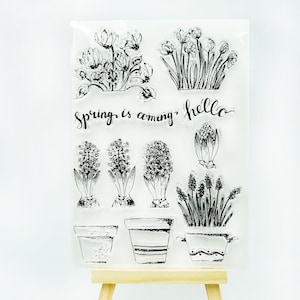 Könnte beinhalten: Klarstempel-Set mit floralen Designs, darunter Topfpflanzen und einzelne Blumen. Der Text "spring is coming" und "hello" sind ebenfalls enthalten. Die Stempel sind in einer Vielzahl von Stilen, ideal für Basteln und Scrapbooking.