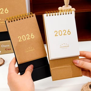 Può includere: Due calendari da scrivania per l'anno 2026. Uno ha una copertina in carta kraft, l'altro una copertina bianca. Entrambi hanno una rilegatura a spirale dorata e l'anno 2026 stampato in oro. Il calendario in carta kraft ha un supporto nero.