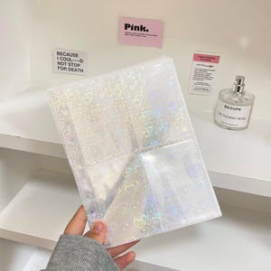 May include: A clear, holographic photo album with heart patterns. The album has multiple pockets for storing photos and is held by a person. The background includes a shelf, stickers that say "Pink" and "BECAUSE I COUL----D NOT STOP FOR DEATH", and a perfume bottle.
