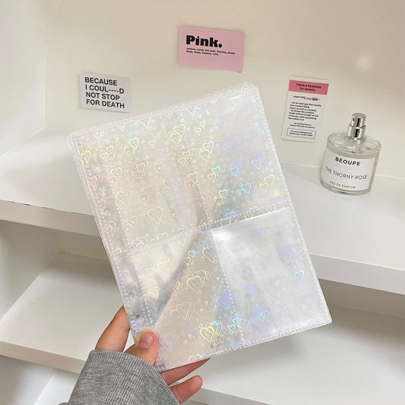 May include: A clear, holographic photo album with heart patterns. The album has multiple pockets for storing photos and is held by a person. The background includes a shelf, stickers that say "Pink" and "BECAUSE I COUL----D NOT STOP FOR DEATH", and a perfume bottle.