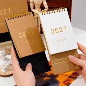 Puede incluir: Dos calendarios de escritorio para el año 2027, uno con cubierta marrón y otro con cubierta blanca. Ambos presentan el texto 2027 y CALENDAR en lámina dorada con cinco estrellas encima. Cada calendario está encuadernado con espiral dorada.