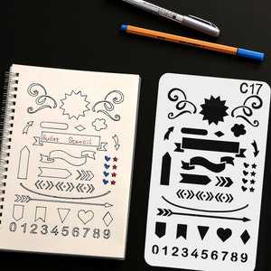 May include: A notebook and stencil for bullet journaling, featuring decorative elements. The notebook has hand-drawn designs including arrows, banners, and numbers. The stencil has similar designs. Pens are visible in the background.