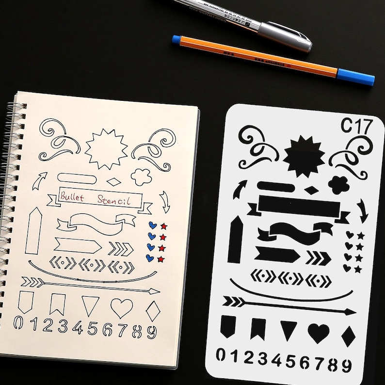 May include: A notebook and stencil for bullet journaling, featuring decorative elements. The notebook has hand-drawn designs including arrows, banners, and numbers. The stencil has similar designs. Pens are visible in the background.
