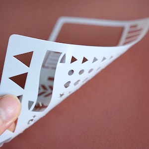 May include: A white plastic stencil with various cut-out shapes, including triangles, circles, and stars. The stencil is held up, revealing its flexibility and the detailed design. The background is a solid, muted reddish-brown.