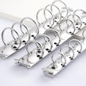 May include: Close-up of several shiny, silver-colored binder rings attached to metal strips. The rings are open and ready to hold paper. The background is white, highlighting the metallic finish of the binder components.