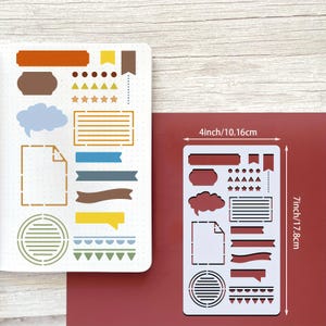 Puede incluir: Una plantilla de pl&aacute;stico blanco con varias formas para journaling, incluyendo banners, bocadillos y estrellas. La plantilla mide 10,16 cm de ancho y 17,8 cm de largo. La plantilla est&aacute; sobre una superficie roja.
