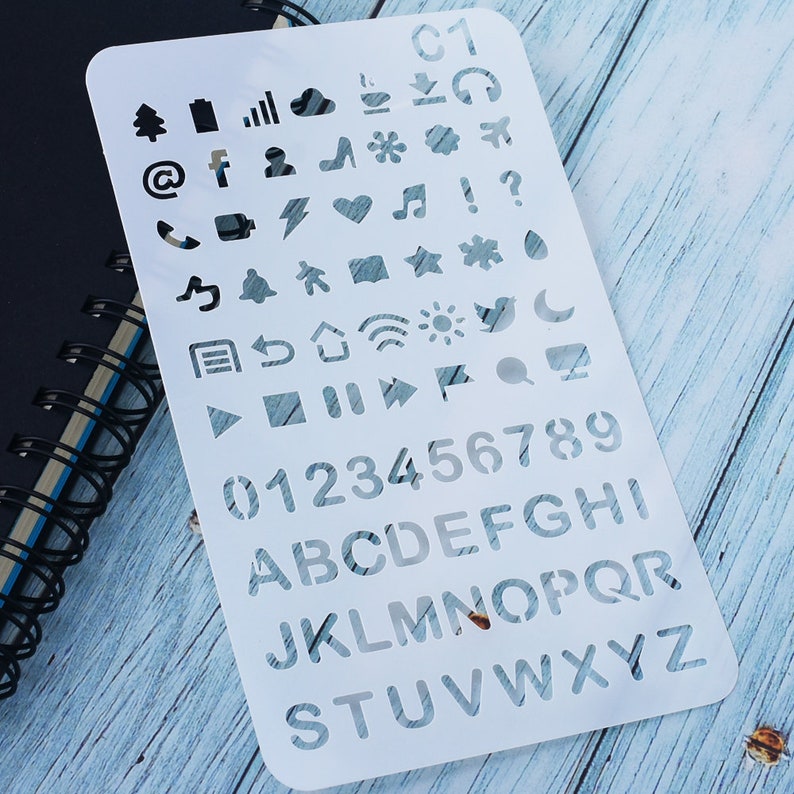 May include: A white plastic stencil featuring cut-out shapes of icons, numbers, and letters. The stencil includes social media symbols, a tree, a bell, and a musical note. Numbers 0-9 and the alphabet are also present.