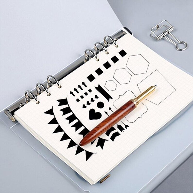 May include: A notebook with a grid pattern, open to a page with black graphic designs, including arrows, hearts, and geometric shapes. A wooden pen with a gold-colored tip rests on the page. A silver binder clip is in the background.