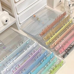 Può includere: Un set di copertine per raccoglitori in plastica trasparente con anelli colorati. Le copertine sono impilate l'una sull'altra e gli anelli sono visibili. Gli anelli sono di diversi colori, tra cui rosa, blu, verde e giallo.