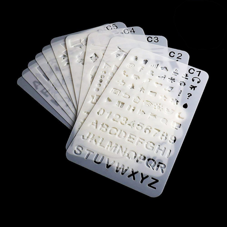 May include: A set of white plastic stencils featuring cut-out letters, numbers, and symbols. The stencils are fanned out, with labels such as C1, C2, C3, C4, and C5 visible. These stencils are designed for various art and craft applications.