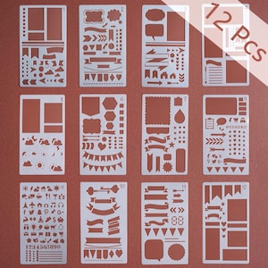 May include: Set of 12 white plastic stencils for bullet journaling and scrapbooking. Each stencil features various shapes, banners, arrows, and icons. The stencils include the text "12 Pcs" and numbers 1-12. The stencils are on a reddish-brown background.