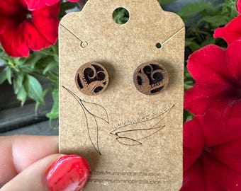 May include: Wooden stud earrings with a dark brown finish and an intricate, carved design. The earrings are displayed on a brown card with the text "Hummingbird Studio" and a leaf illustration. The background features vibrant red flowers.
