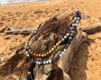 Könnte beinhalten: Zwei Perlenketten mit blauen, beigen und hellblauen Perlen, die auf einem Stück Treibholz an einem Sandstrand liegen. Eine marineblaue Quaste mit goldenen Akzenten hängt an der unteren Halskette. Das Meer ist im Hintergrund zu sehen.
