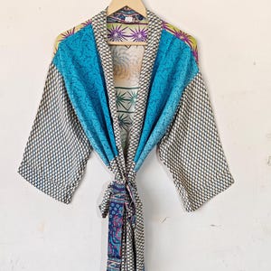 May include: A colorful, patterned robe with a tie closure, hanging against a white wall. The robe features a mix of blue, white, and patterned fabrics. The sleeves have a geometric design, and the front has a solid blue panel.