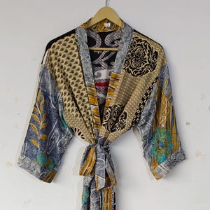 May include: A patchwork robe with a tie belt, hanging on a wooden hanger. The robe features a mix of patterns and colors, including beige, blue, and black. The sleeves and body are made of different fabric panels.