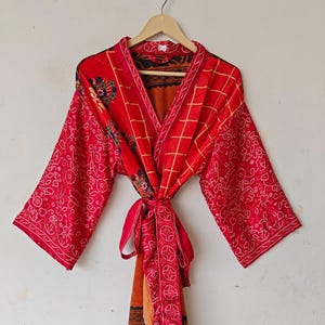 May include: A striking red robe with a wrap-around style, showcasing a variety of patterns. The robe has long sleeves with a dotted design and a tie belt. The interior is a warm orange colour, and it hangs on a wooden hanger.