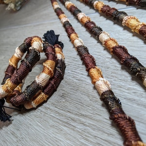 May include: Four long strands of braided hair extensions in brown, beige, and black colors. The strands are twisted together and have a tassel at the end.