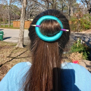 Könnte beinhalten: Ein türkisfarbener Schaumstoff-Haarkissen mit einem violetten und hölzernen Haarstift, der durch ihn hindurchgeht und einen Dutt aus braunem Haar hält.