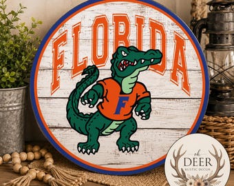 May include: A round wooden sign with a white faux-plank background featuring the word FLORIDA in large orange letters and a green alligator mascot wearing an orange shirt with a blue F. The sign has a thick blue and orange circular border.