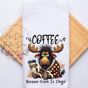 May include: White waffle-weave kitchen towel with a cartoon moose wearing a brown robe and holding a coffee cup. The towel has the word "COFFEE" and the phrase "Because Crack Is Illegal" printed on it. The towel is on a wooden cutting board.