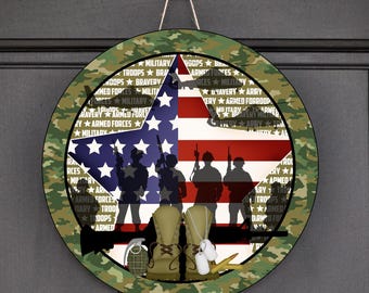 Op de afbeelding: Ronde houten deurhanger met een camouflage rand. Het ontwerp toont een Amerikaanse vlag met silhouetten van soldaten, laarzen, hondenpenningen en een granaat. De woorden "Military", "Troops" en "Bravery" zijn rond de vlag gedrukt.