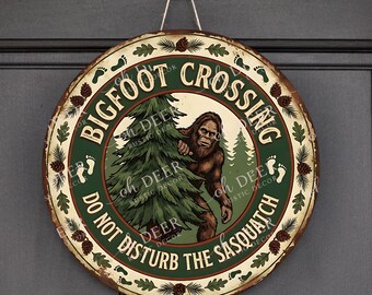 Könnte beinhalten: Ein rundes Metallschild mit einer braunen Bigfoot-Illustration, die hinter einem grünen Tannenbaum hervorkommt. Der Rand ist cremefarben mit Tannenzapfen und Fußabdrücken. Der Text lautet BIGFOOT CROSSING und DO NOT DISTURB THE SASQUATCH.