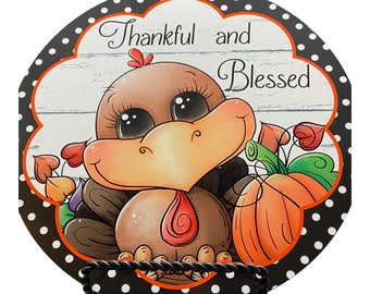 Pode incluir: Um sinal redondo de madeira com fundo de bolinhas pretas e brancas. O sinal apresenta um peru de desenho animado com uma abóbora e o texto "Thankful and Blessed".