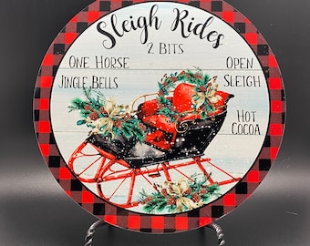Könnte beinhalten: Ein rundes, rot-schwarzes kariertes Schild mit dem Text "Sleigh Rides 2 Bits" und Bildern eines Schlittens, eines Pferdes und Glöckchen. Das Schild zeigt auch "One Horse Jingle Bells", "Open Sleigh" und "Hot Cocoa".