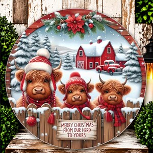 May include: A round decorative sign featuring three Highland cattle wearing winter hats and scarves behind a wooden fence. The scene includes a red barn, a red truck, and snow-covered pine trees. A sign on the fence reads, Merry Christmas from our herd to yours.