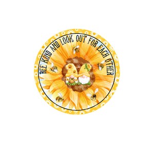 Op de afbeelding: Een gele zonnebloem met twee dwergen in het midden. De zonnebloem heeft een witte rand met kleine witte bloemen. De tekst "Bee Kind and Look Out for Each Other" staat rond de zonnebloem.