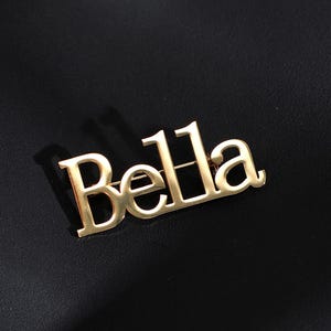 May include: A gold-colored pin with the name "Bella" in a stylized font. The pin is set against a black background, creating a contrast that highlights the lettering. The pin is likely a fashion accessory.