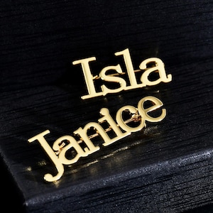 May include: Two gold-colored metal name pins, one with the name "Isla" and the other with the name "Janice".