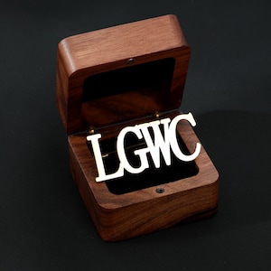 May include: A wooden jewelry box with a hinged lid, open to reveal a gold-colored metal inscription that reads "LGWC". The box is lined with black velvet and sits against a dark background. The wood has a rich, dark brown color.