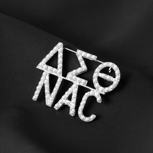 May include: Silver-toned brooch featuring the Greek letters Delta, Sigma, Theta, and the letters NAC, all encrusted with small, round, white pearls. The brooch is set against a black background.