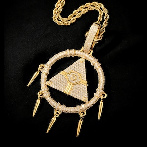 Puede incluir: Collar con colgante dorado con un triángulo dentro de un círculo, incrustado con pequeñas piedras brillantes. El colgante tiene dijes colgantes en forma de púas. La cadena es de diseño de cuerda trenzada. Fondo negro.
