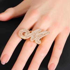 May include: A gold-toned ring in the shape of the letter "M", encrusted with clear, sparkling crystals. The ring is a statement piece, with a large, ornate design. The crystals are set closely together, creating a dazzling effect. The ring is worn on a finger.