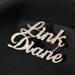 Könnte beinhalten: Eine goldfarbene Metallbrosche mit den Namen Link und Diane in Schreibschrift. Die Oberfläche der Buchstaben ist mit kleinen, klaren, funkelnden Strasssteinen besetzt, die das Licht einfangen und für ein helles, reflektierendes Aussehen sorgen.