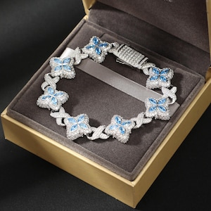 May include: A silver bracelet with blue gemstone flower-shaped details, displayed in a gold box with a grey interior. The bracelet features a series of interconnected floral designs, each with a central blue stone and surrounding clear stones.