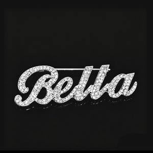 Puede incluir: Broche plateado que deletrea "Bella" en una fuente cursiva. Las letras están incrustadas con cristales transparentes y brillantes. El broche tiene un cierre de barra clásico en la parte posterior, sobre un fondo negro.