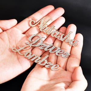 Può includere: Tre spille per nome dorate con decorazioni in strass trasparenti. I nomi sono "Natasha", "Amber" e "Divine".